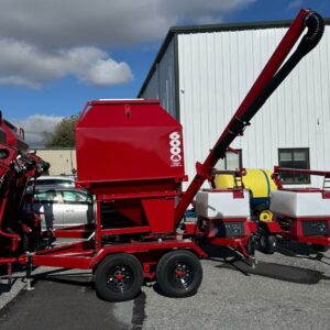 The 3-Ton Mobile Bulk Feed Bin 6000 with Solar-Powered Auger, painted red with a tall conveyor, stands on a paved lot beside a white metal building under a partly cloudy sky.